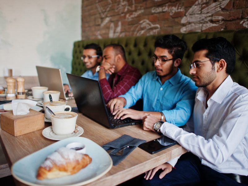 Group of four south asian men's posed at business meeting in cafe. Indians work with laptops together using various gadgets, having conversation.